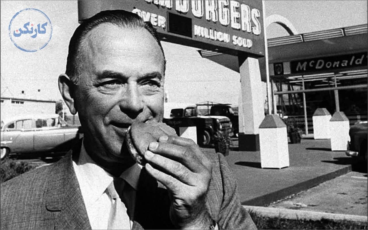 عکس سیاه سفید از ری کراک موسس مک دونالد در حالی که با لبخند در حال خوردن همبرگر است و در پشت نمایی از رستوران مک دونالد دیده می‌شود.