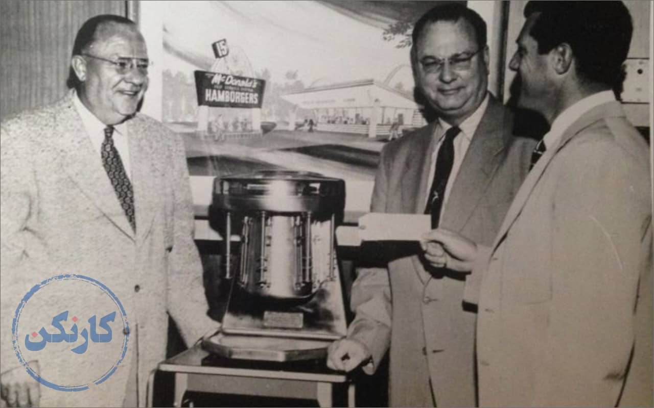 عکس قدیمی سیاه سفید از ری کراک موسس مک دونالد درحال دادن چک به برادران مک دونالد که در کنار یک دستگاه مالتی میکسر ایستاده اند